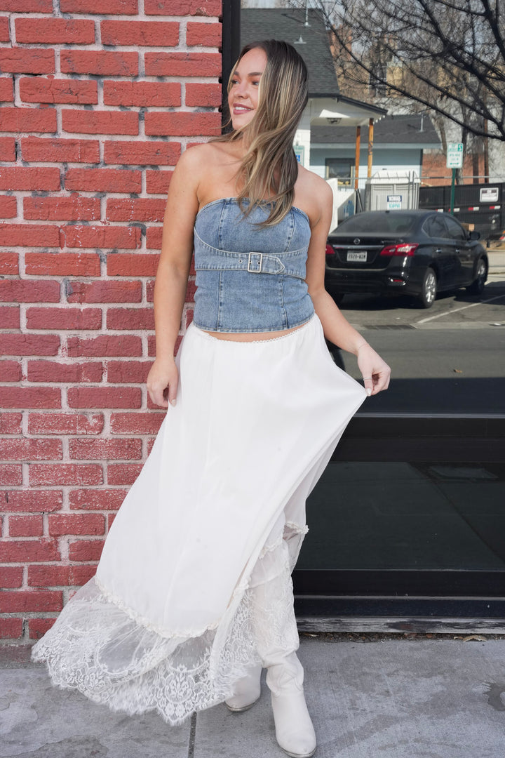 Woman wearing a denim strapless top and white lace skirt standing against a brick wall.