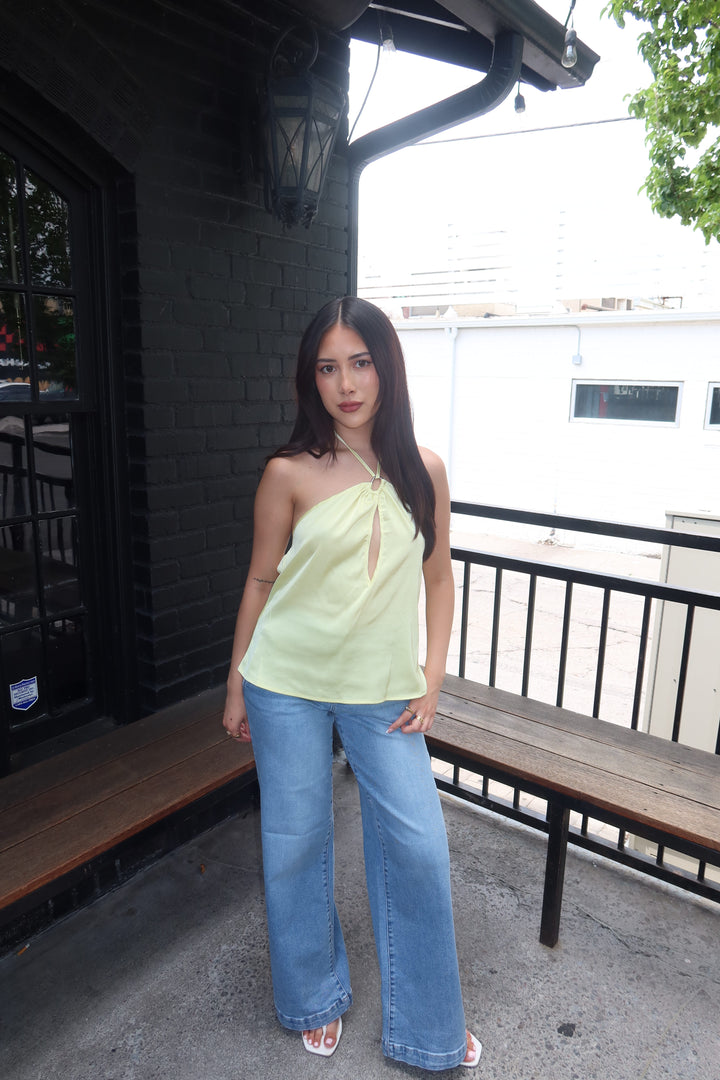 A woman stands on a porch wearing a pale yellow-green halter top with a keyhole cutout and light-wash wide-leg jeans.