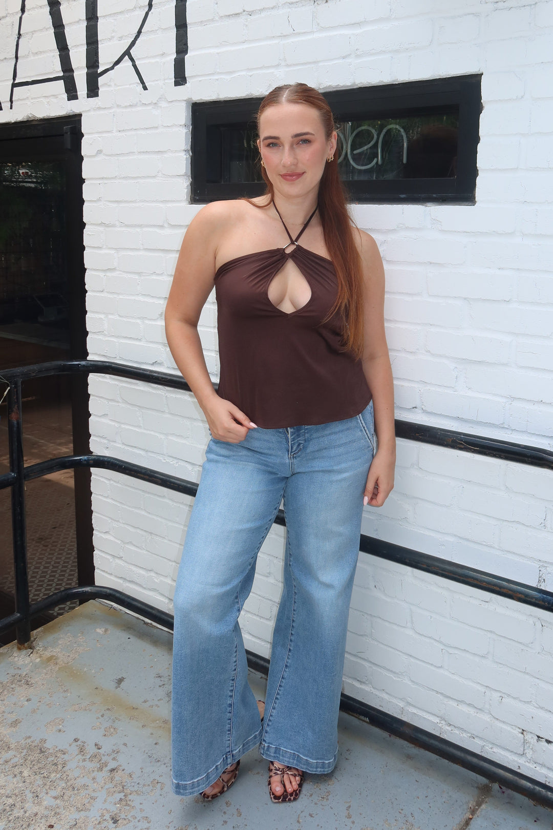 A woman stands outdoors against a white brick wall wearing a dark brown halter-style cutout top and light blue wide-leg jeans.
