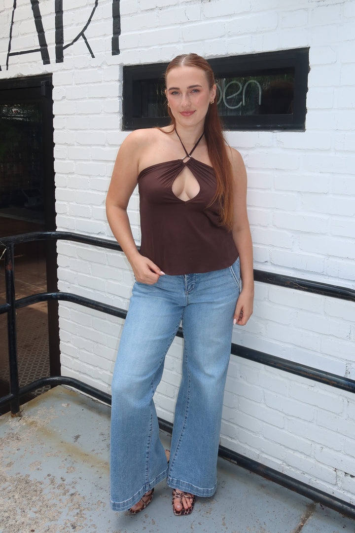 A woman stands outdoors against a white brick wall wearing a dark brown halter-style cutout top and light blue wide-leg jeans.