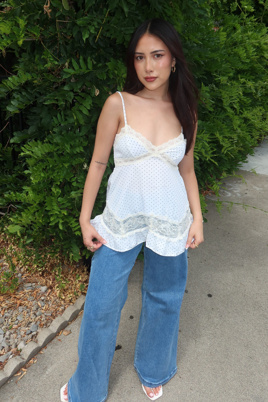 A woman stands outdoors against a green hedge wearing a white lace-trimmed camisole top with wide-leg blue jeans and white sandals.