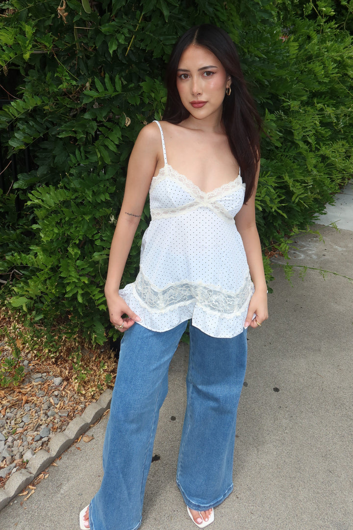 A woman stands outdoors against a green hedge wearing a white lace-trimmed camisole top with wide-leg blue jeans and white sandals.