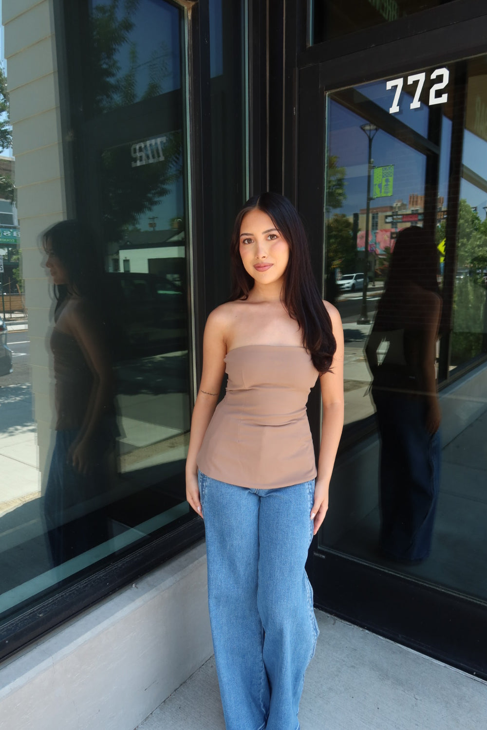 A woman stands outdoors in front of a storefront window wearing a strapless beige top and light blue jeans. The building has large black-framed glass windows, and the number 772 is visible on the door.