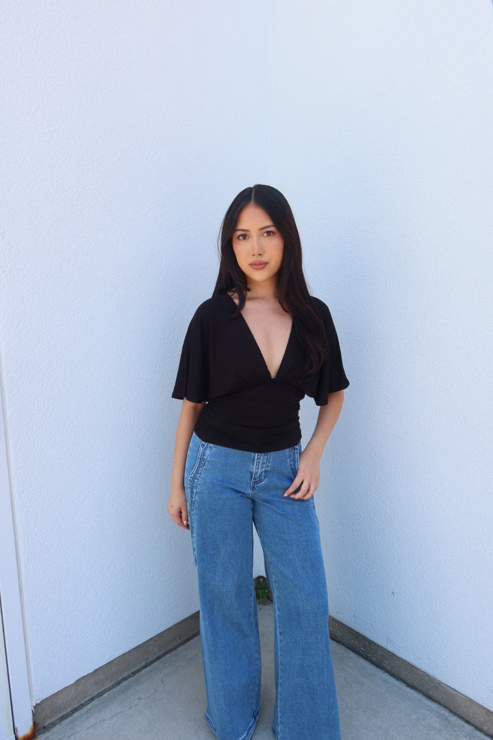 A woman with long dark hair stands against a light blue wall wearing a black short-sleeve wrap-style top and wide-leg blue jeans.