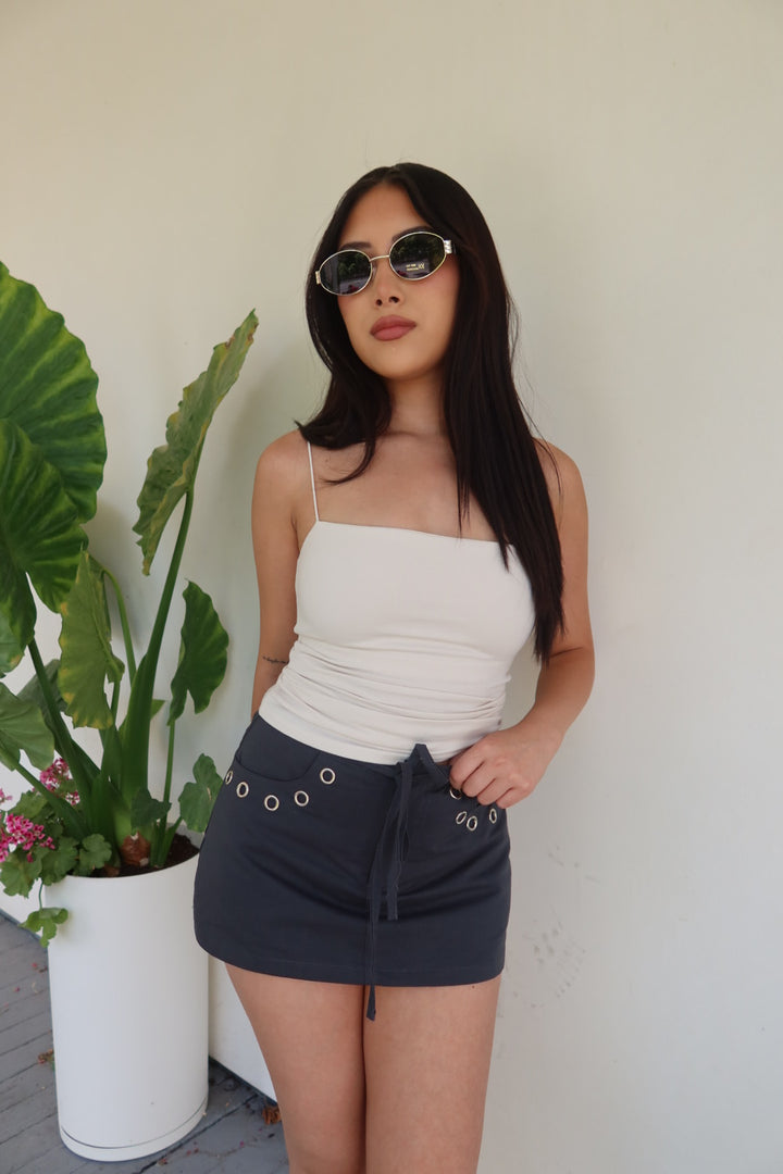 A woman with long dark hair and sunglasses stands against a white wall wearing a white ribbed camisole and a dark gray mini skirt with silver grommets. A large green potted plant is on the left.