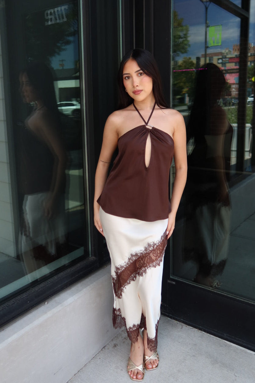 A woman models a two-piece dress with a brown halter top and a white skirt featuring a brown lace-like pattern, standing in front of a building entrance with glass doors.