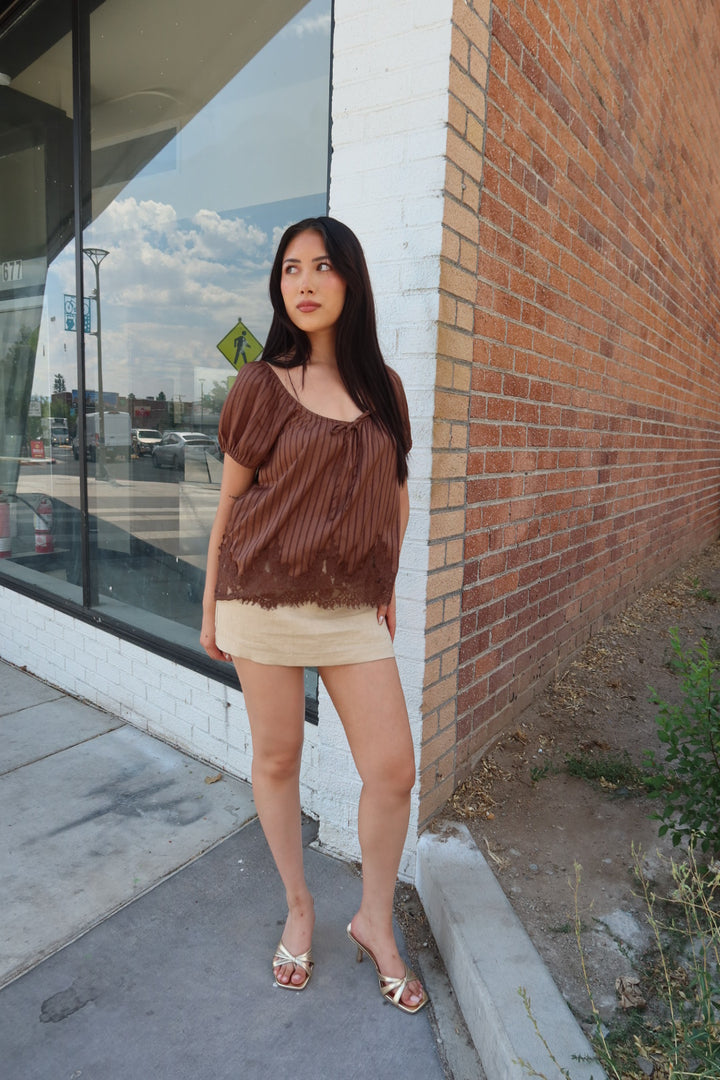 A woman stands outdoors on a sidewalk beside a brick wall and storefront window, wearing a brown pleated short-sleeve top with a frayed hem and a beige mini skirt, paired with metallic heeled sandals.
