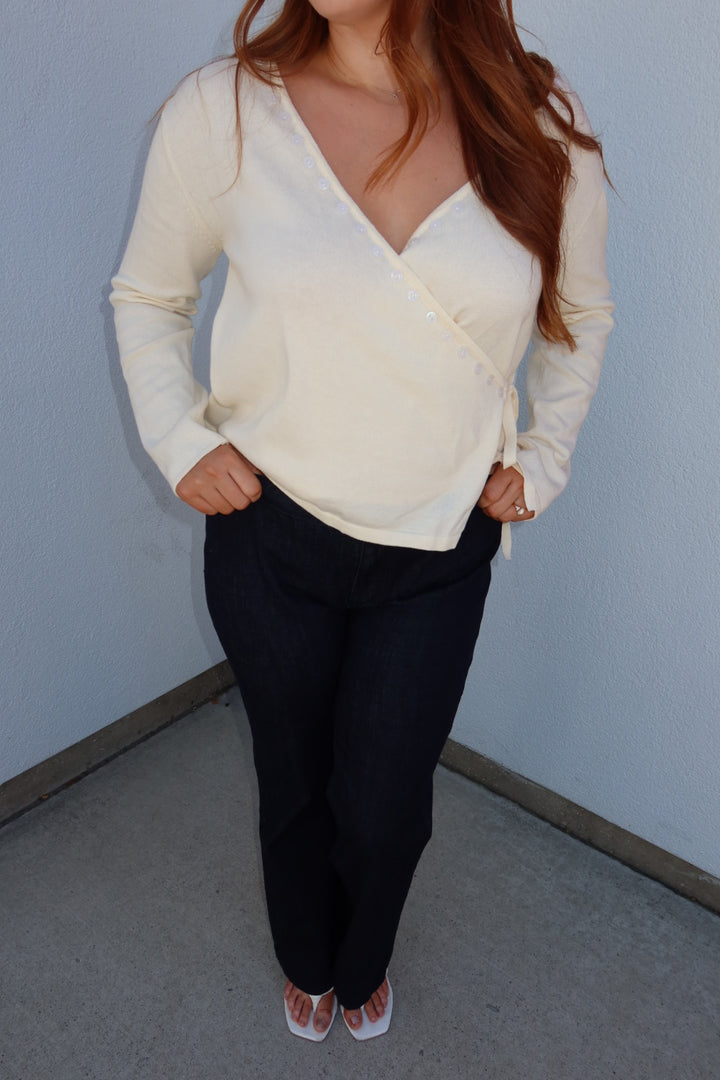 A woman wearing a cream-colored long-sleeve wrap-style knit top and dark navy pants, standing in a corner against a light blue-gray wall.