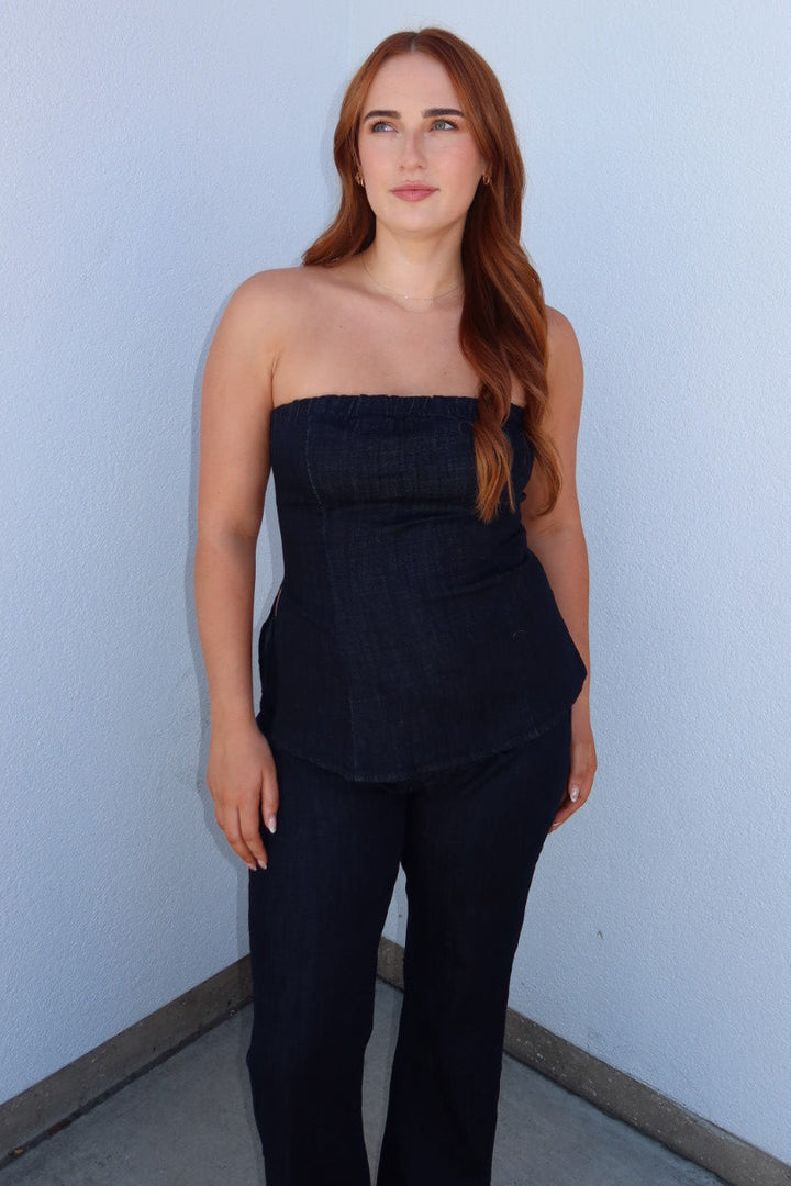 A woman with long red hair stands in a corner wearing a dark navy strapless top and matching dark pants against a white wall with a red accent.