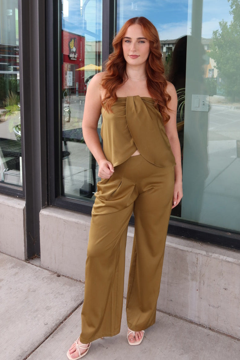 A woman with long red hair stands outdoors in front of a building with large glass windows, wearing a sleeveless olive-brown jumpsuit and light pink sandals.