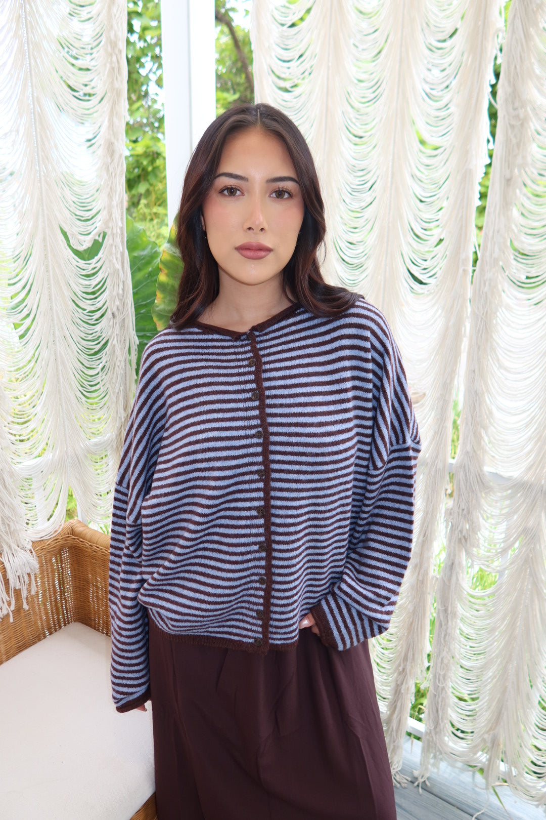 A woman wearing a navy and white striped long-sleeve knit top with brown buttons, paired with a dark brown skirt, standing in front of white macrame curtains.