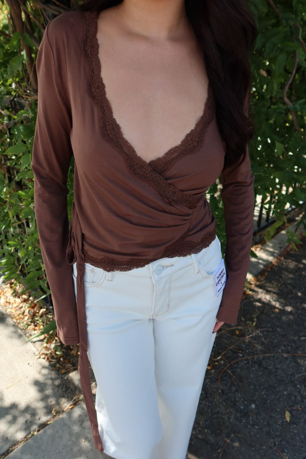 A woman wearing a brown long-sleeve wrap-style top with lace trim and white pants, standing outdoors in front of green foliage.