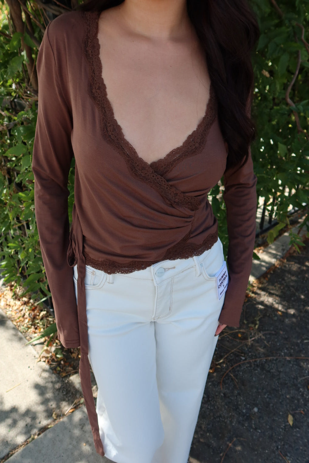 A woman wearing a brown long-sleeve wrap-style top with lace trim and white pants, standing outdoors in front of green foliage.