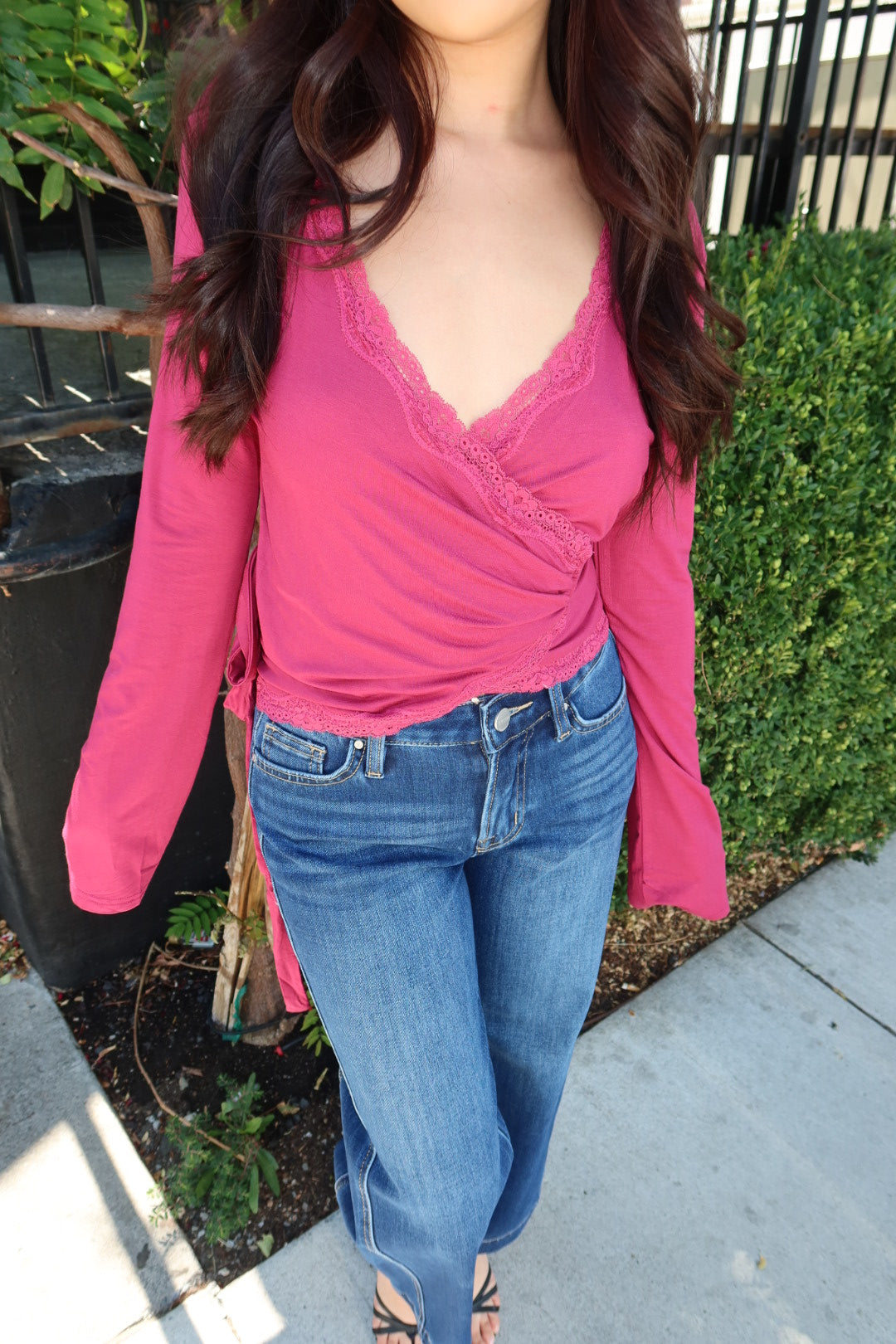 A woman wearing a bright pink long-sleeve wrap-style top with lace trim and blue jeans, standing outdoors on a sidewalk next to green hedges.