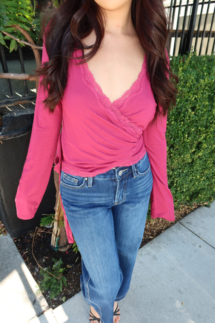 A woman wearing a bright pink long-sleeve wrap-style top with lace trim and blue jeans, standing outdoors on a sidewalk next to green hedges.