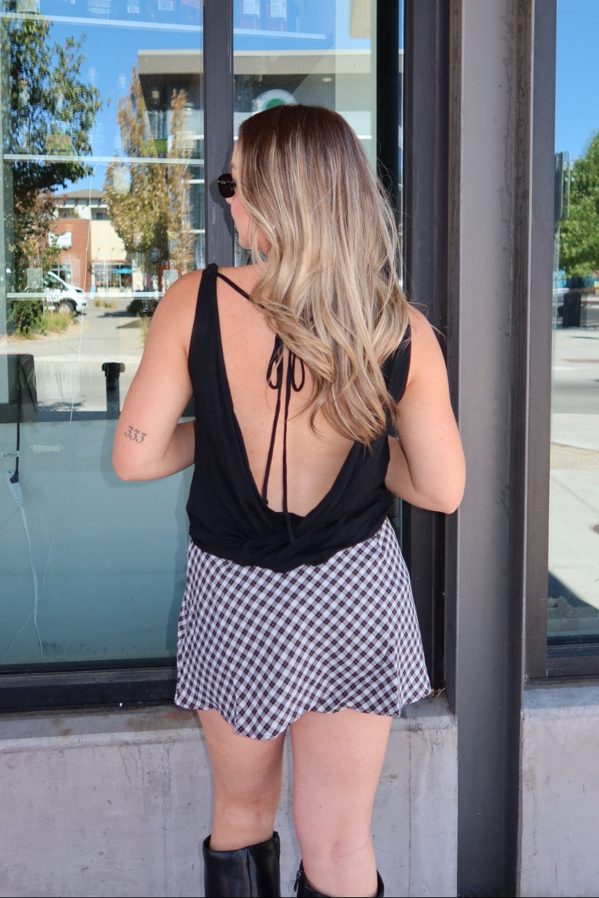 A woman viewed from behind wearing a black back-tie tank top and a black and white gingham mini skirt, standing in front of a storefront window.