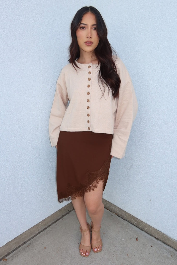 Model in the brown Lucy asymmetric lace trim satin midi skirt and a cropped cream cardigan, full length front view