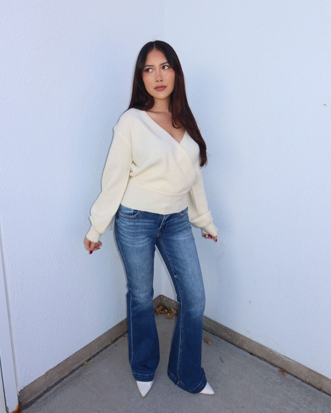 A woman stands in a corner wearing a cream-colored wrap-style knit sweater, blue flared jeans, and white pointed-toe heels against a pale blue wall.
