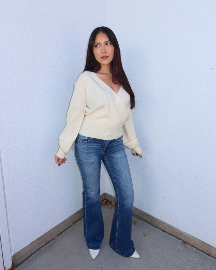 A woman stands in a corner wearing a cream-colored wrap-style knit sweater, blue flared jeans, and white pointed-toe heels against a pale blue wall.
