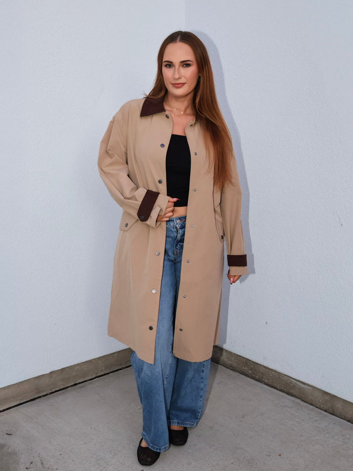 Woman wearing a beige trench coat, black top, and blue jeans against a white wall.
