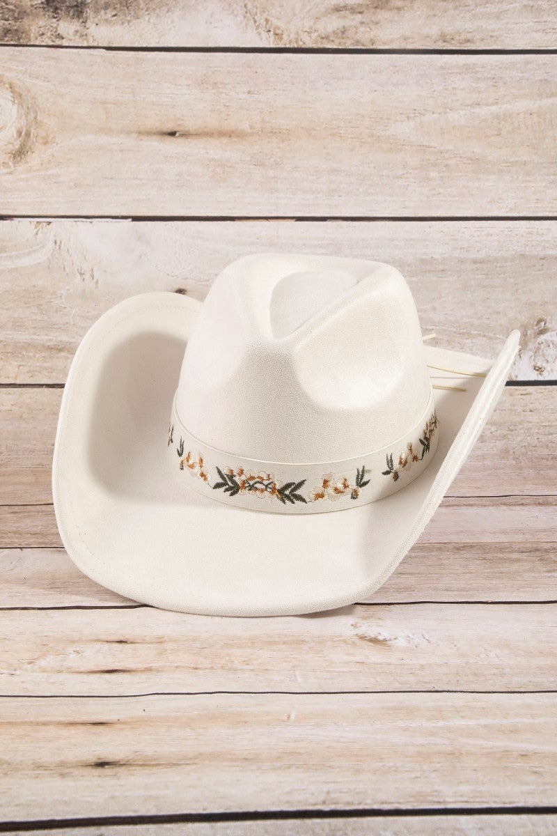 The Botanical Embroidered Strap Cowboy Hat in White - Three-Quarter View on White Wood Surface Showing Crisp Ivory Felt Body, Wide Brim & Sage Green and Gold Botanical Embroidered Hatband