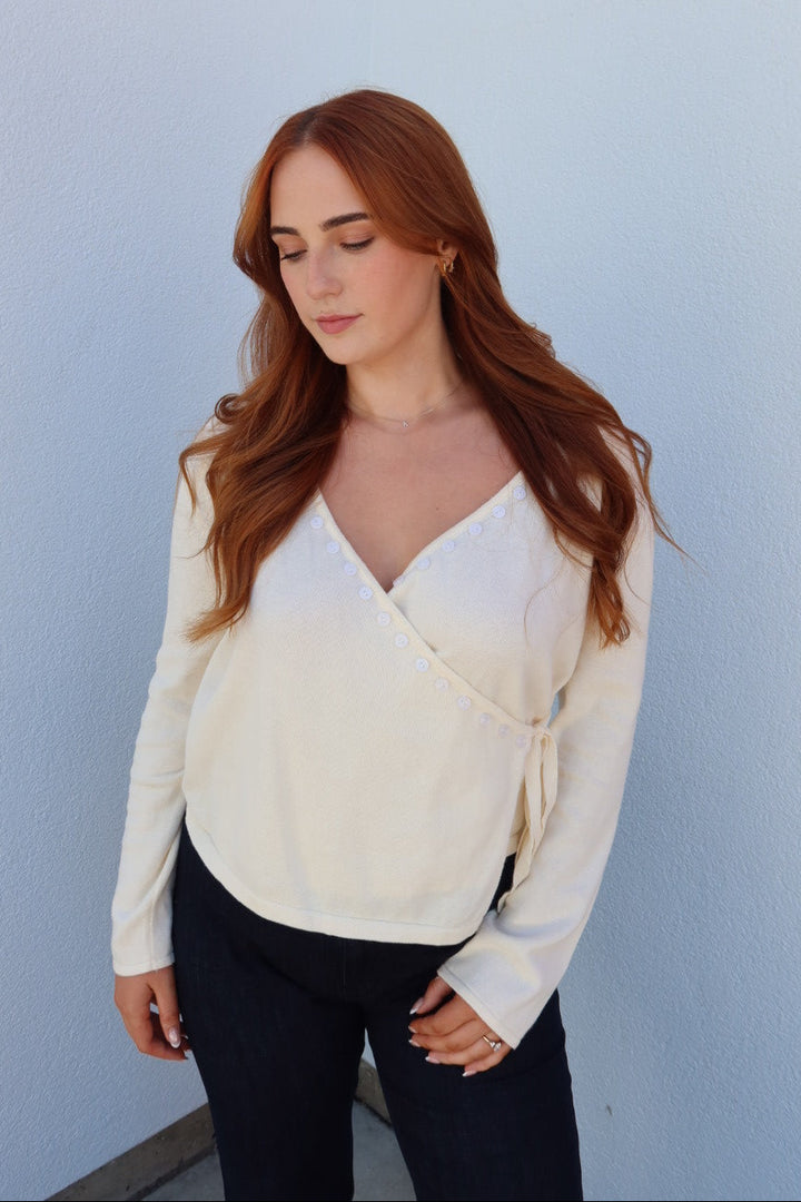 A woman with long red hair wearing a cream-colored long-sleeve wrap top and dark pants, standing against a light blue wall.