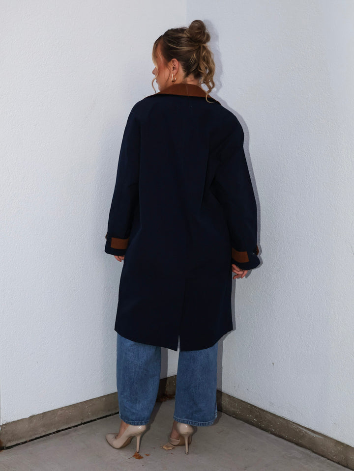 Person wearing a dark coat and blue jeans standing against a white wall.