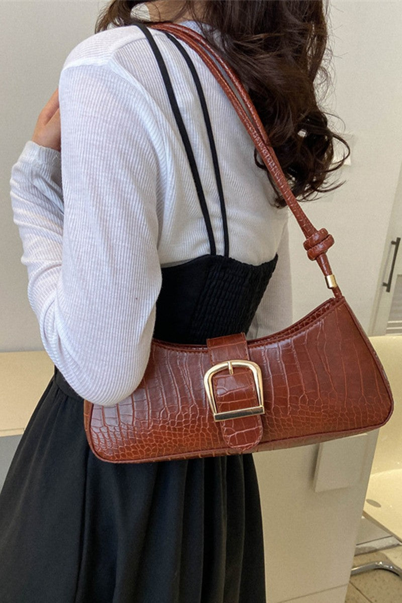 A woman wearing a white ribbed long-sleeve top and black skirt carries a brown crocodile-embossed shoulder bag with a gold-tone buckle strap.