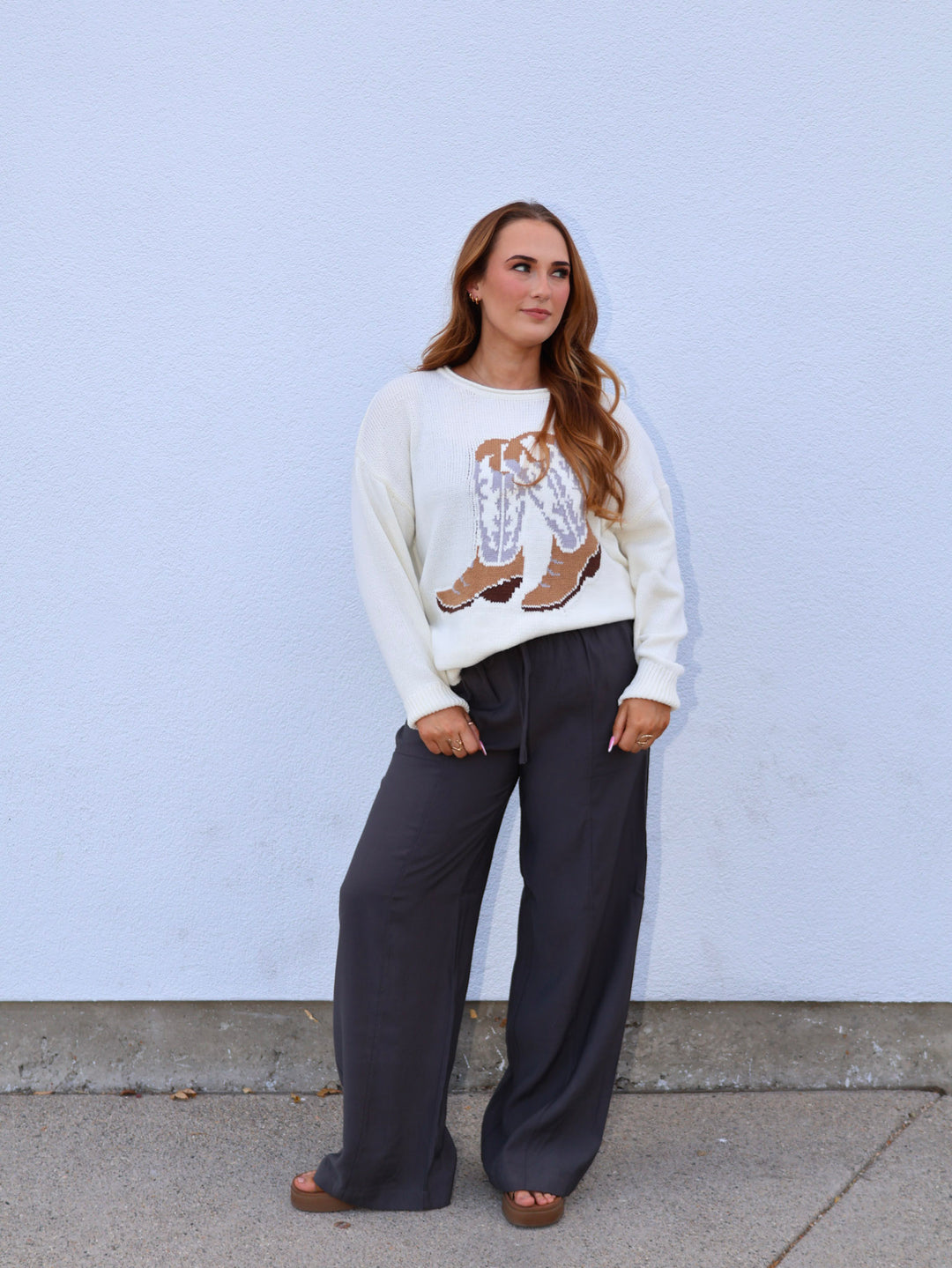 Woman wearing a white sweater with a graphic design and dark pants against a light blue wall.