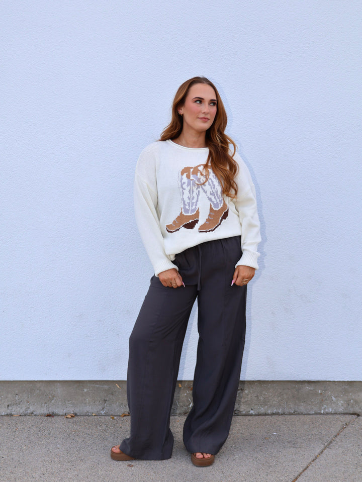 Woman wearing a white sweater with a graphic design and dark pants against a light blue wall.