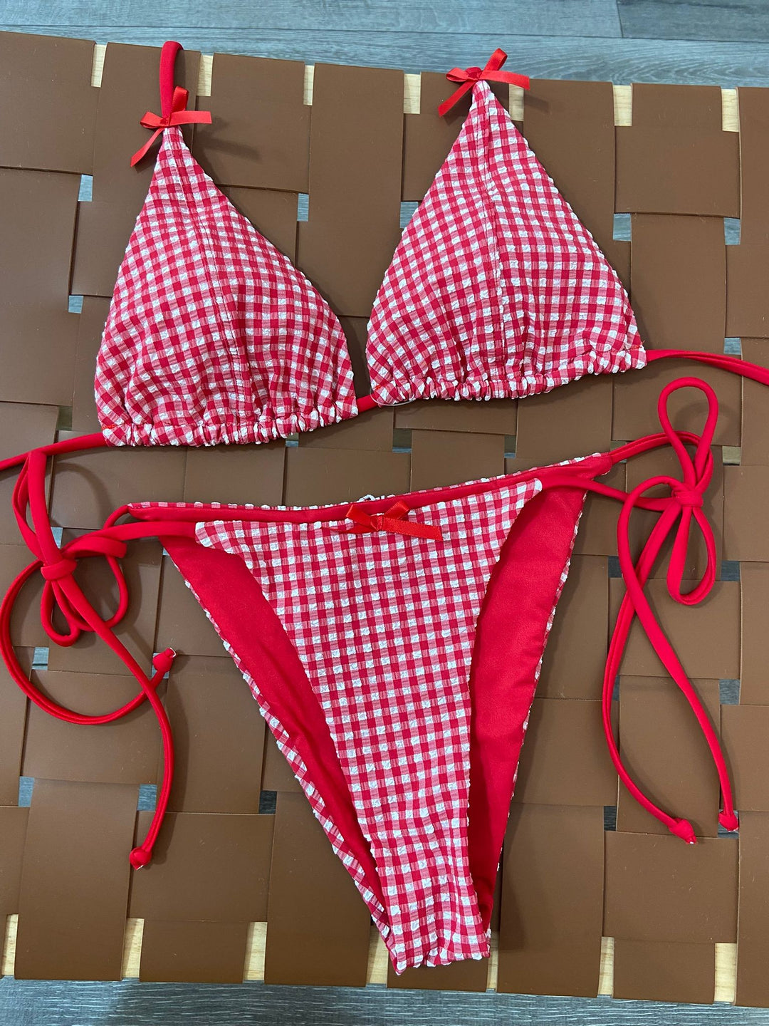 The Cherry Gingham Triangle Bikini Set - Flat Lay on Rattan Surface Showing Full Set Detail — Gingham Seersucker Texture, Bow Tie Details & Red Side Tie Bottoms