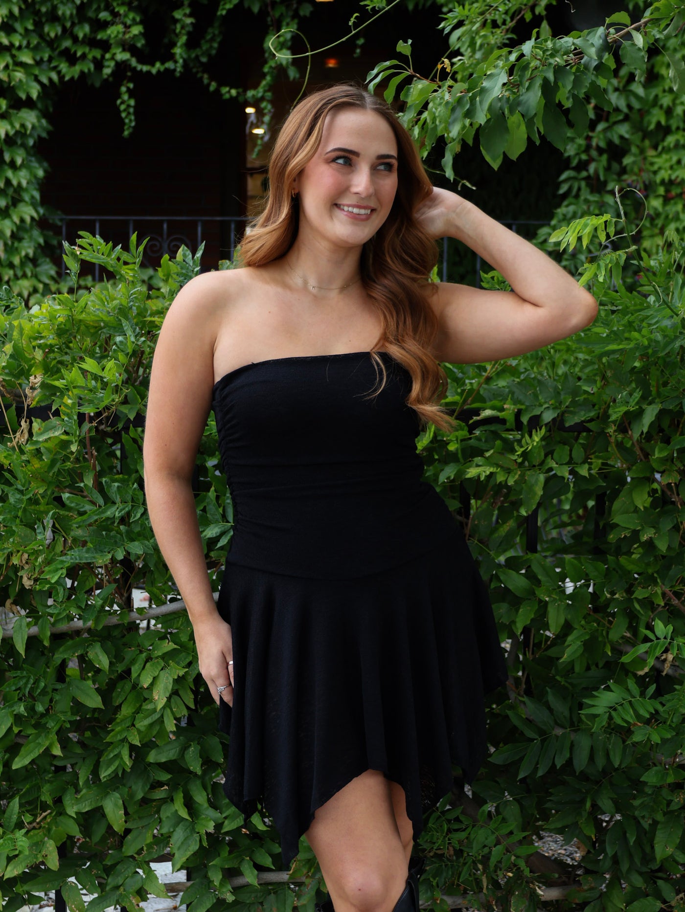 Woman wearing a black strapless dress standing in front of green foliage.