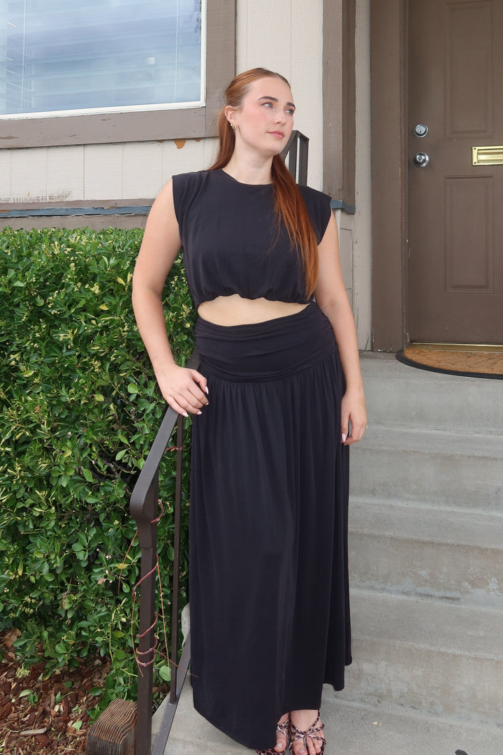 A woman with red hair stands on the concrete steps of a house wearing a sleeveless black crop top and a long black skirt.