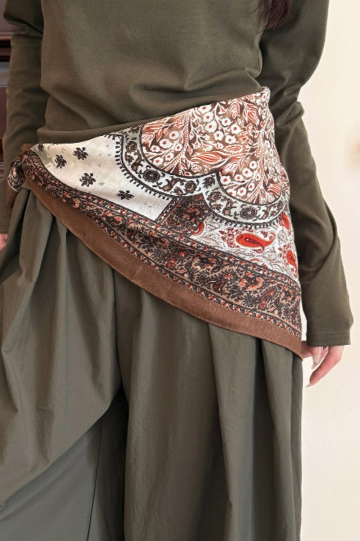 A close-up of a person wearing an olive green outfit with a patterned scarf wrapped around the waist. The scarf features a cream base with brown, rust, and black paisley and floral motifs.