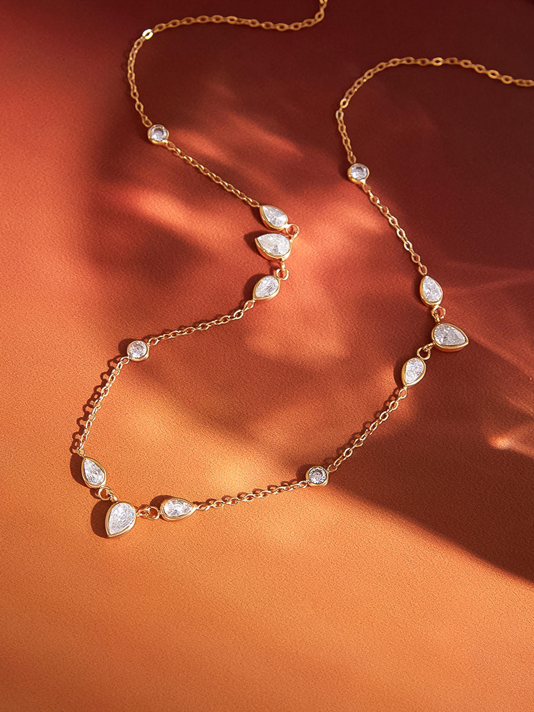 The CZ Teardrop Station Layering Necklace in 18K Gold - Artistic Flat Lay on Warm Terracotta Background in Dappled Light Showing Full Chain with Alternating Round & Teardrop CZ Bezel Stations