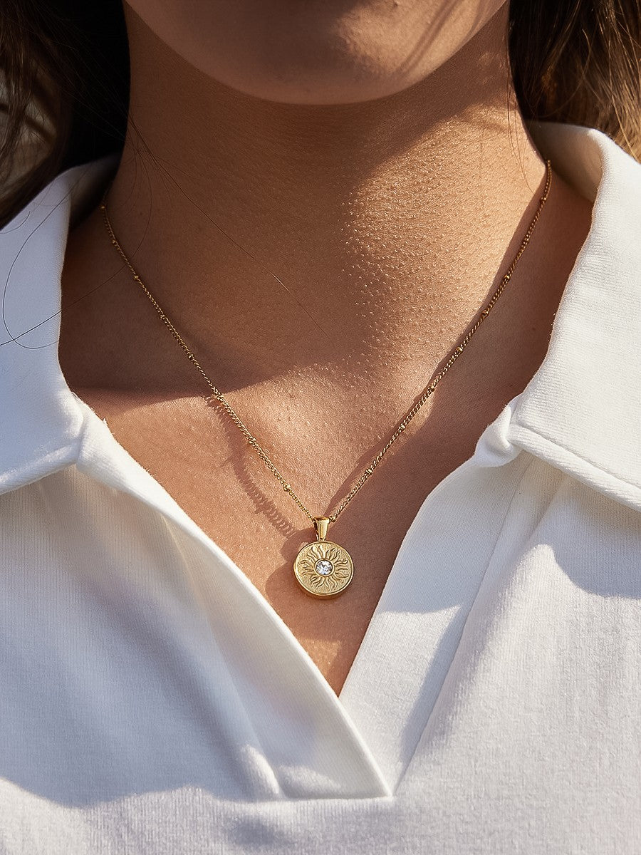 The CZ Sun Medallion Coin Necklace in 18K Gold - Outdoor On-Model Lifestyle Shot Showing Sun Medallion Coin Pendant on Satellite Chain at Collarbone in Natural Sunlight Against White Linen Shirt