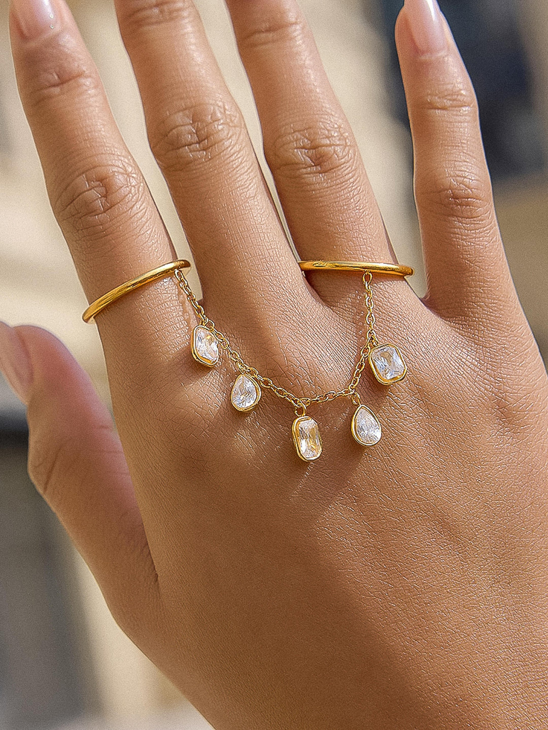 The CZ Dangle Chain Double Ring in 18K Gold - On-Model Close-Up on Hand Against Warm Background Showing Both Gold Bands on Index & Middle Fingers with Five CZ Dangle Charms Hanging from Chain