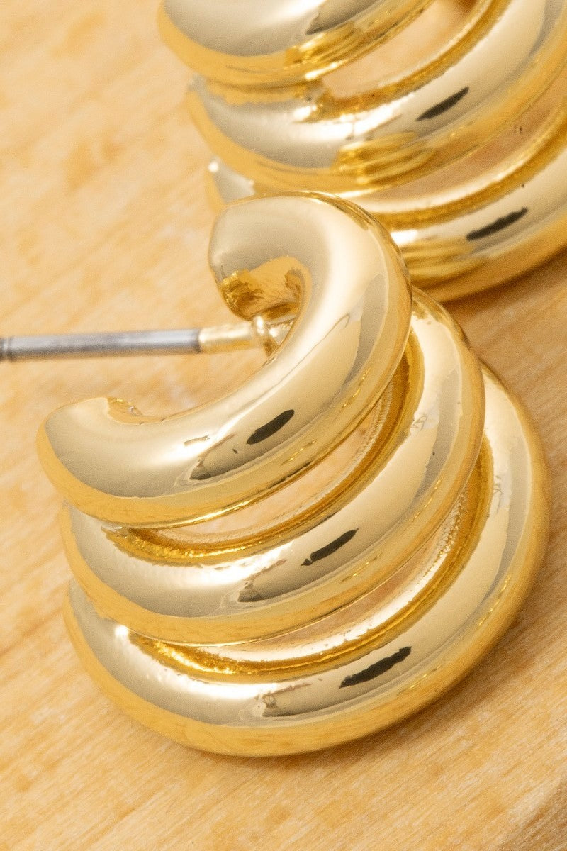 The Columbus Triple Coil Hoop Earrings in 18K Gold Dipped - Close-Up Detail Shot on Wood Surface Showing Spiraling Triple Coil Formation, Polished Gold Finish & Post Back Closure