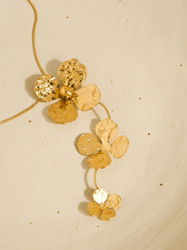 The Triple Hammered Flower Lariat Necklace in 18K Gold - Product Flat Lay on Speckled Cream Background Showing Full Cascade of Three Hammered Gold Flower Pendants on Double Snake Chain with Hammered Texture Detail