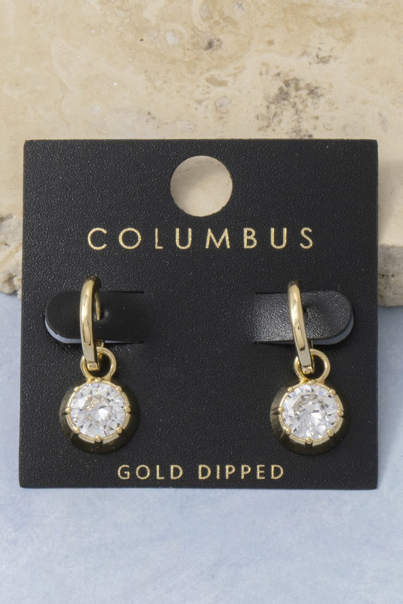 The Columbus CZ Bezel Charm Huggie Earrings in Gold Dipped - Product Shot on Black Columbus Card Against Stone Background Showing Pair of Gold Huggie Hoops with Round CZ Bezel Charm Drops