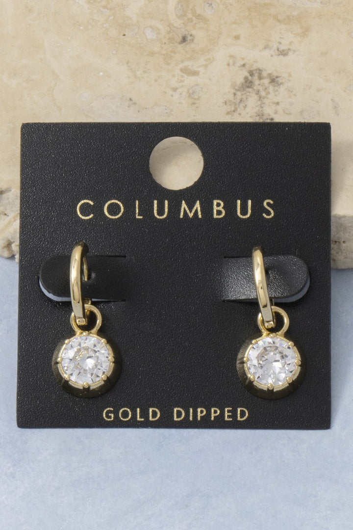The Columbus CZ Bezel Charm Huggie Earrings in Gold Dipped - Product Shot on Black Columbus Card Against Stone Background Showing Pair of Gold Huggie Hoops with Round CZ Bezel Charm Drops