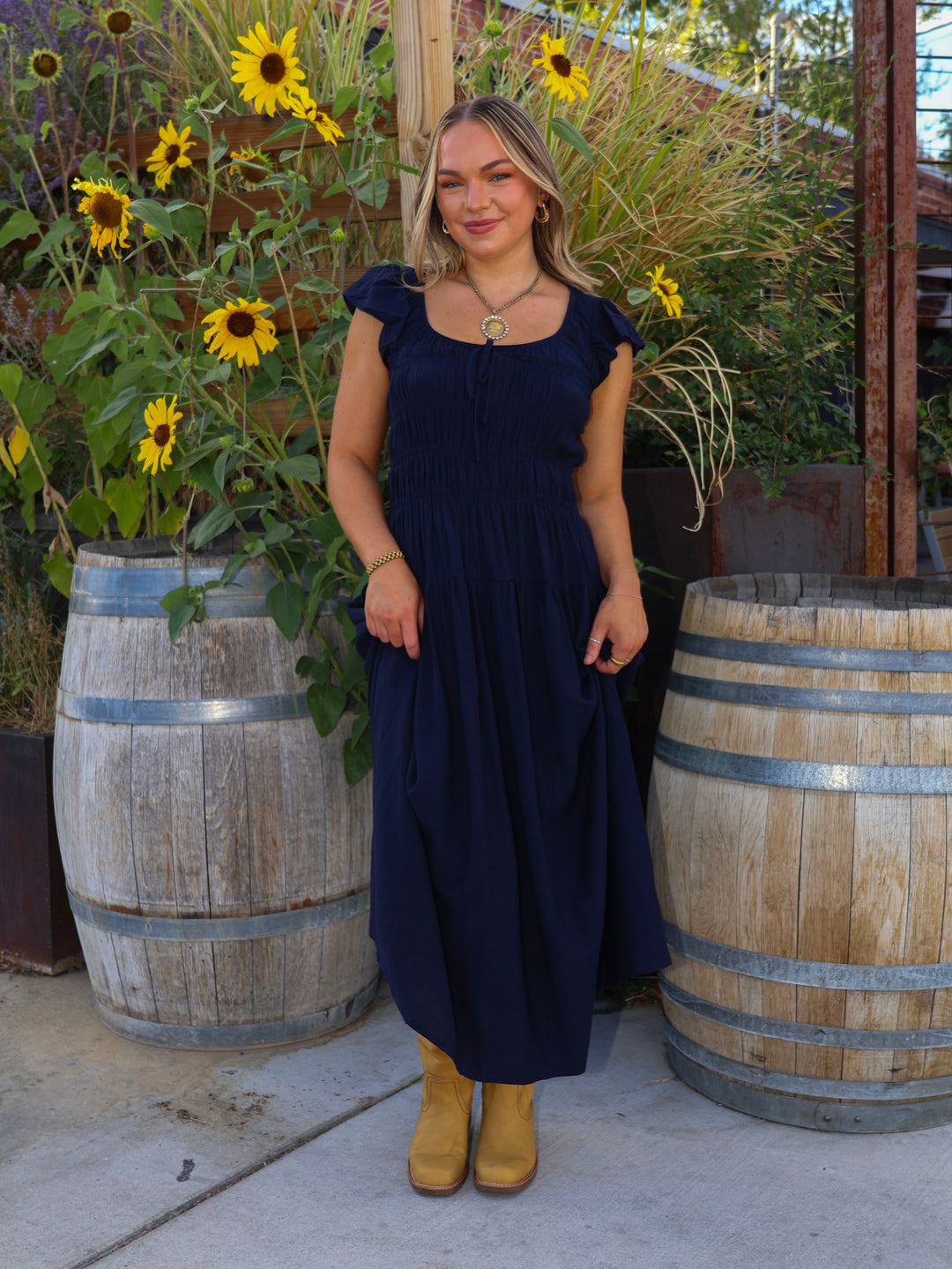 Woman in a navy blue dress standing in front of sunflowers and wooden barrels.