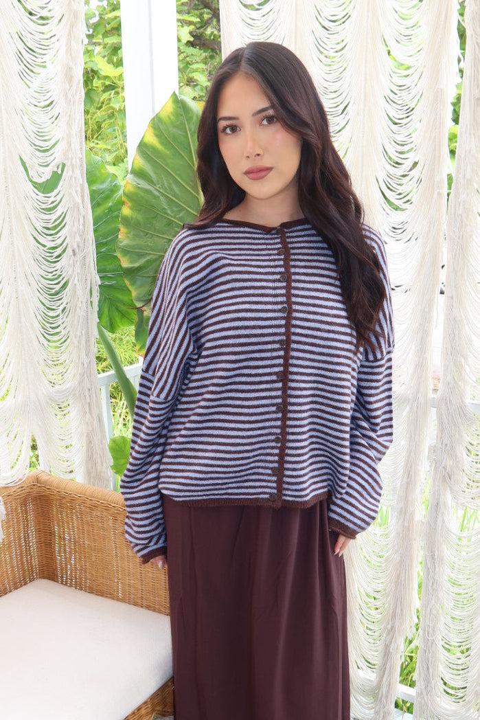 A woman stands on a porch wearing a navy and white striped long-sleeve top with brown trim and a dark brown maxi skirt. The background features white macrame curtains and a wooden bench.