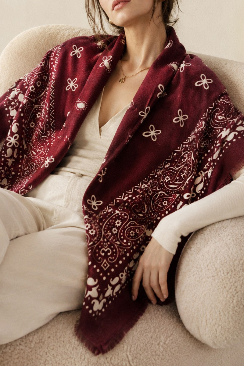 A woman seated on a cream-colored sofa wearing a burgundy shawl with white paisley and floral motifs over a cream top and cream pants.