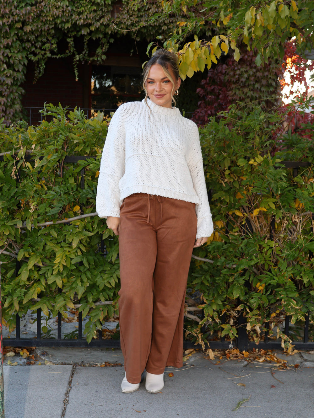 Woman wearing a white sweater and brown pants standing in front of green bushes.