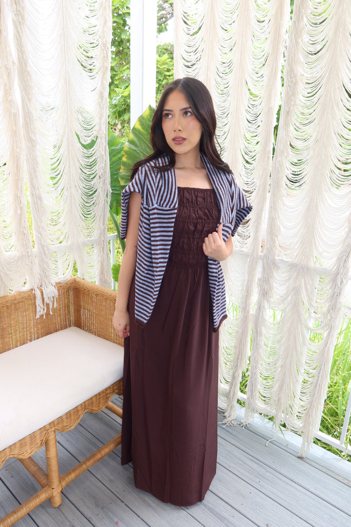 A woman stands on a porch wearing a long dark brown dress with a blue and white striped cardigan. The background features white macrame wall hangings and a wicker bench.
