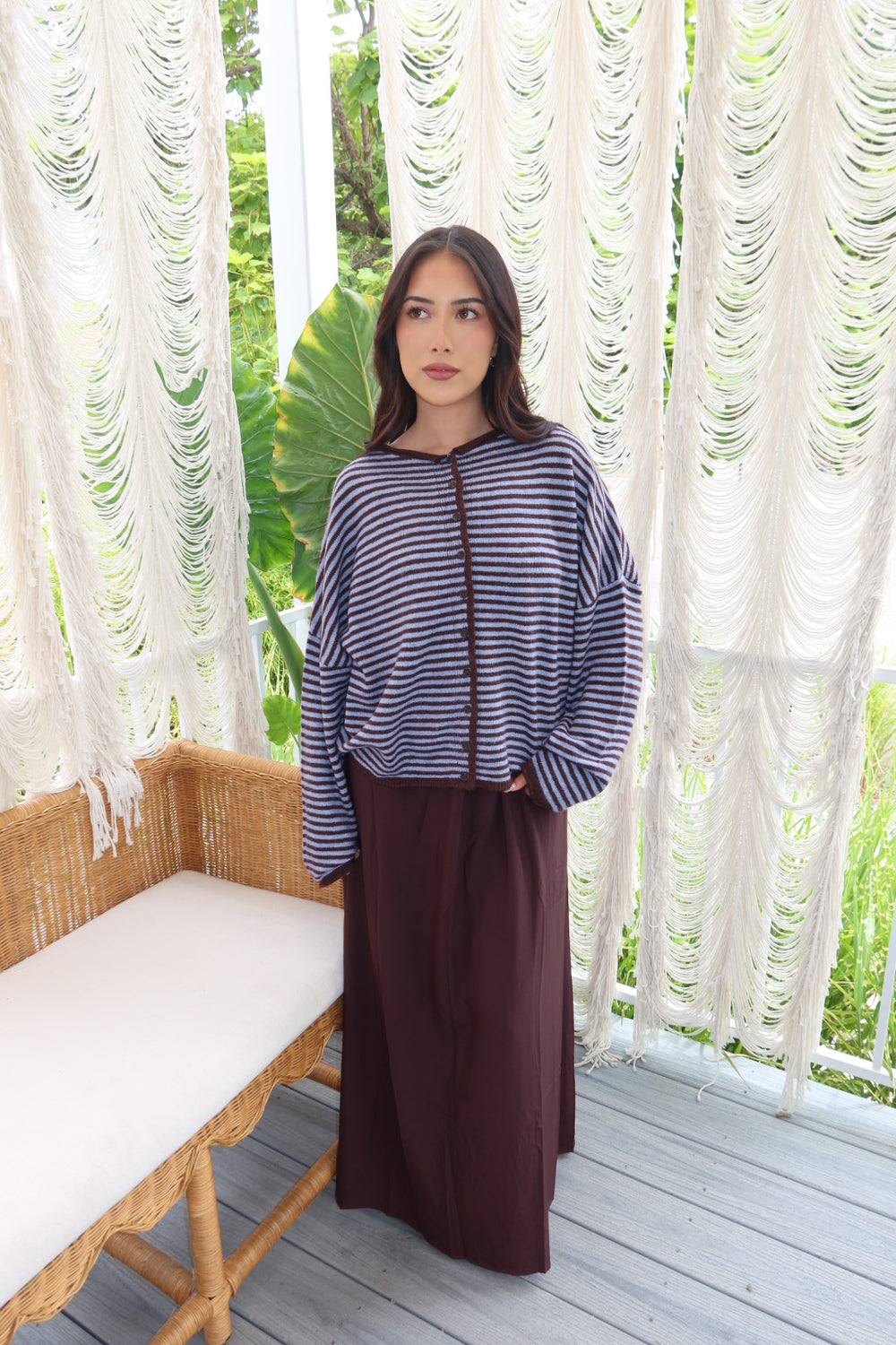 A woman stands on a porch wearing a navy and white striped long-sleeve top with a brown cord detail and a dark brown skirt. The background features white macrame curtains and a wicker bench.