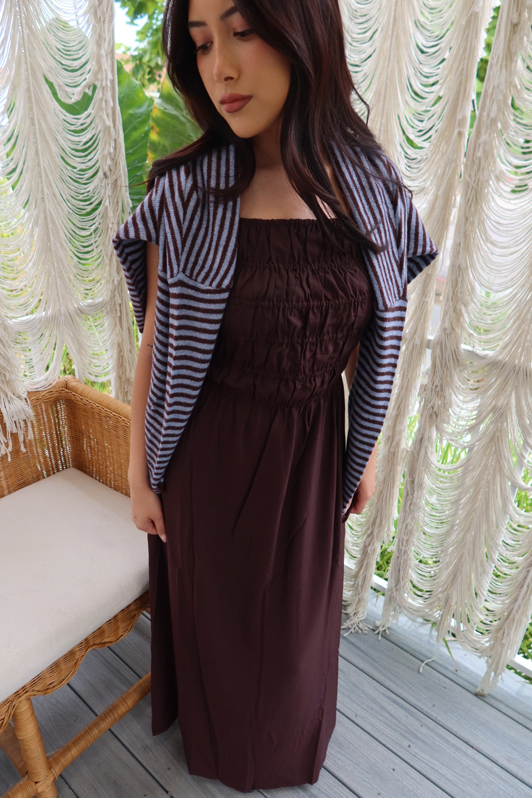 A woman wearing a dark brown smocked maxi dress with a navy and white striped cardigan, standing on a wooden deck in front of white macrame curtains.