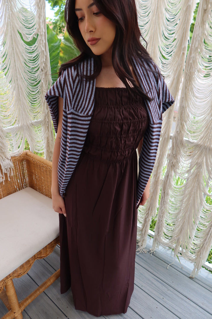 A woman wearing a dark brown smocked maxi dress with a navy and white striped cardigan, standing on a wooden deck in front of white macrame curtains.