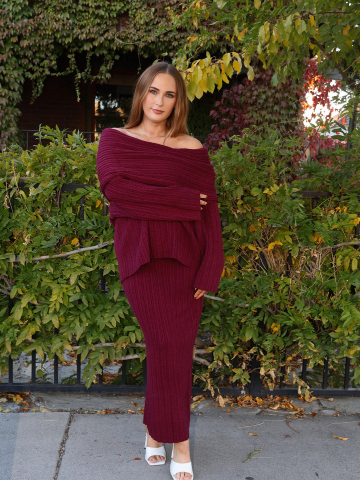 Woman wearing a burgundy off-shoulder top and skirt standing in front of green bushes.